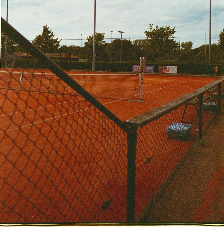 A scenic view of an outdoor clay tennis court with net and fencing on a sunny day.