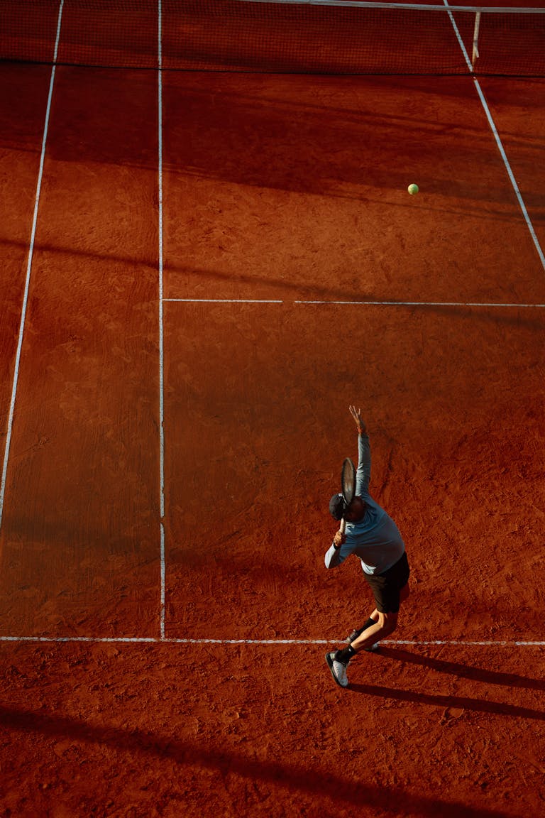 Aerial view of a tennis player serving on a clay court during a match at dusk.