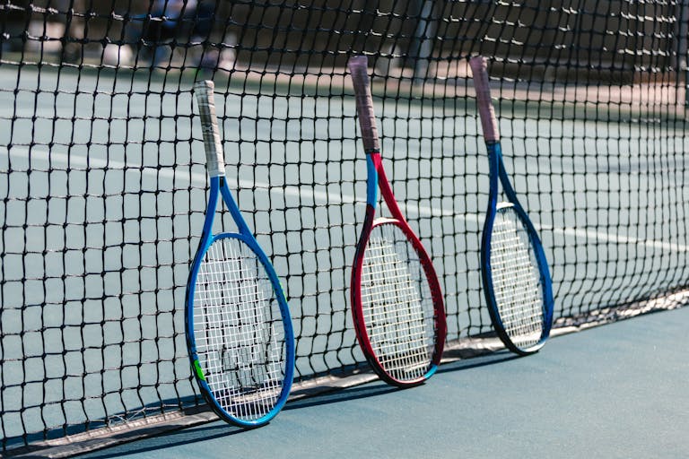 Three colorful tennis rackets leaning against a net on an outdoor tennis court.