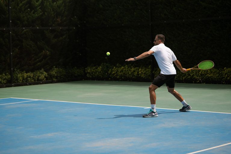 Tennis player hitting a backhand shot on a sunny outdoor court in Iran.